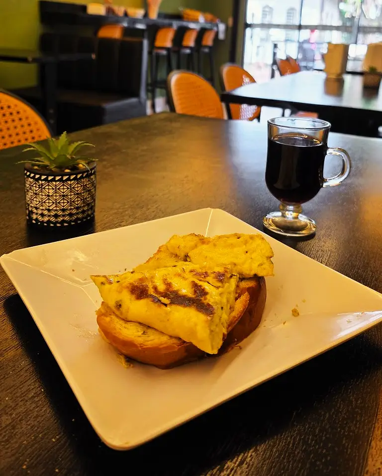 Pão na chapa com omelete em prato branco, ao lado de um café preto